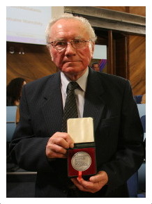 Portret mężczyzny w okularach, ubranego w ciemny garnitur, białą koszulę i krawat. Mężczyzna trzyma przed sobą otwarte, czerwone etui, w którym znajduje się srebrny medal. Postać stoi w pomieszczeniu z drewnianą boazerią w tle, obok fragmentu ekranu projekcyjnego.