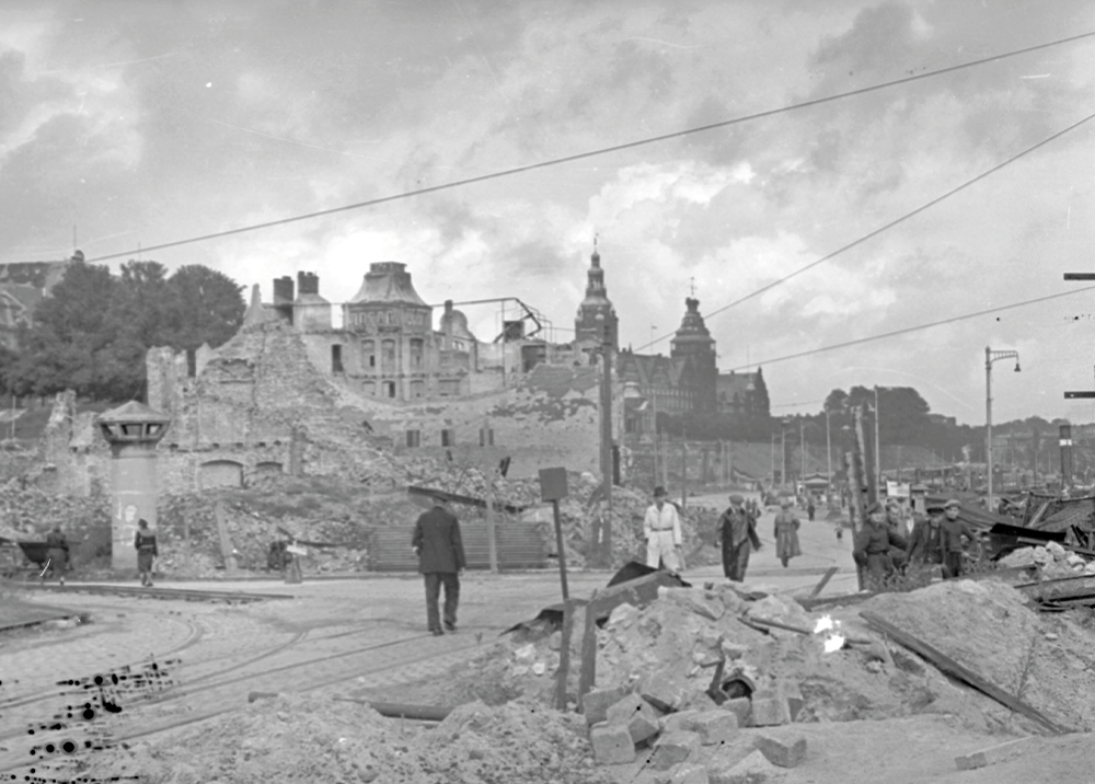 Czarno-biała fotografia dokumentalna z okresu bezpośrednio po II wojnie światowej, przedstawiająca zniszczone centrum Szczecina. Widok z perspektywy ulicy wypełnionej stertami gruzu, cegieł i materiałów budowlanych na pierwszym planie. Po lewej stronie widać tory tramwajowe przecinające drogę, a obok nich betonowy słup ogłoszeniowy lub budkę strażniczą. W centralnej części kadru znajduje się duża, częściowo zniszczona ruina okazałego budynku, prawdopodobnie dawnej kamienicy lub pałacu, z widocznymi odsłoniętymi wnętrzami i szkieletem dachu. W oddali widać panoramę zrujnowanego miasta z charakterystycznymi wieżami kościołów (w tym Katedry św. Jakuba) i innych budynków, rozpoznawalnych jako Szczecin (np. Wały Chrobrego, Czerwony Ratusz). Kilka postaci ludzkich widać idących po rumowisku, w tym mężczyzna w płaszczu. Nad ulicą rozciągnięte są liczne kable i przewody telegraficzne na słupach.