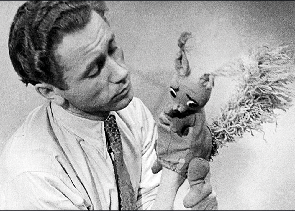 Czarno-biała fotografia portretowa średniego planu przedstawiająca Stanisława Laguna jako młodego mężczyznę. Patrzy czule w lewo, trzymając uniesione ręce, na których umieszczona jest ręczna pacynka w kształcie wiewiórki. Lagun ma na sobie jasną koszulę z ciemnym, wzorzystym krawatem. Pacynka-wiewiórka, wykonana z futrzastego materiału, ma duże, wyraziste oczy i puszysty ogon zakręcony za głową, trzymając uniesioną łapkę. Relacja wzrokowa między mężczyzną a lalką sugeruje komunikację. Tło jest proste, jasne i niewyraźne.