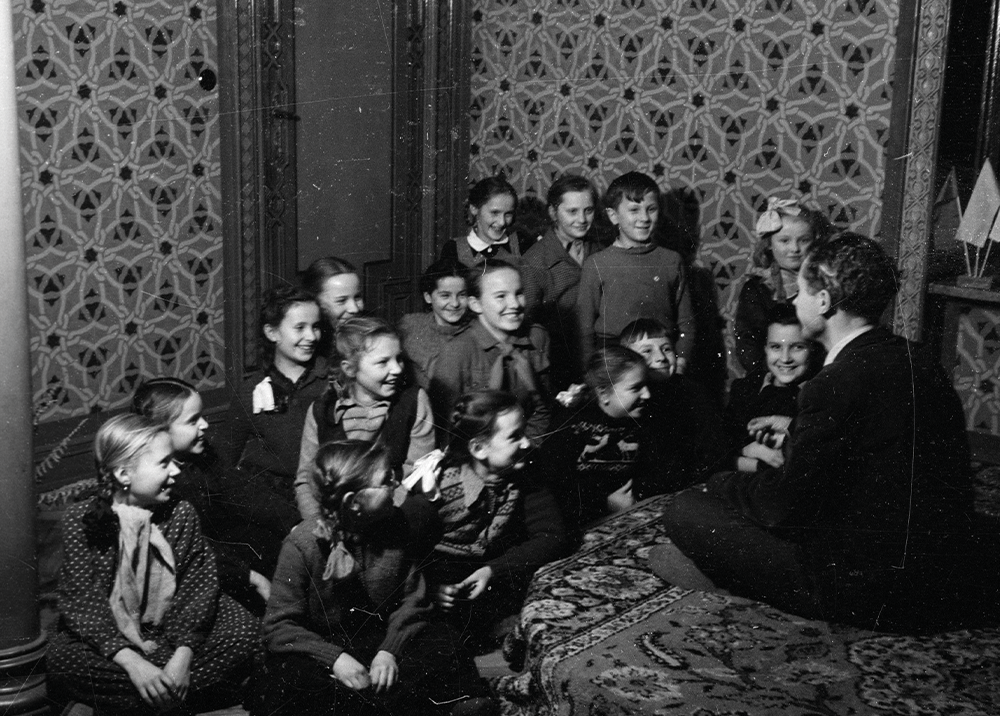 Archiwalna, czarno-biała fotografia przedstawiająca grupę osób siedzących na wzorzystym dywanie i podłodze wewnątrz bogato zdobionego pomieszczenia. Wszyscy zgromadzeni patrzą z uśmiechem i uwagą na mężczyznę siedzącego tyłem do aparatu, który gestykuluje podczas rozmowy. Ściany pokoju pokryte są tapetą o gęstym, geometrycznym wzorze, a w tle widać elementy ozdobnej stolarki.