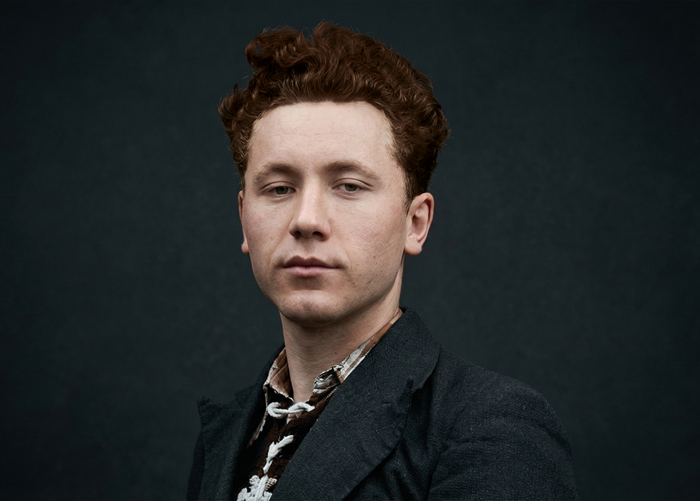 Portret młodego Stanisława Laguna na jednolitym, czarnym tle. Mężczyzna o poważnym wyrazie twarzy i gęstych, ciemnych, falowanych włosach, ubrany w klasyczny strój z połowy XX wieku: ciemną marynarkę o wyraźnej strukturze, kamizelkę w drobną kratkę oraz jasną koszulę w cienkie, pionowe paski. Światło miękko pada na twarz, podkreślając historyczny charakter fotografii.