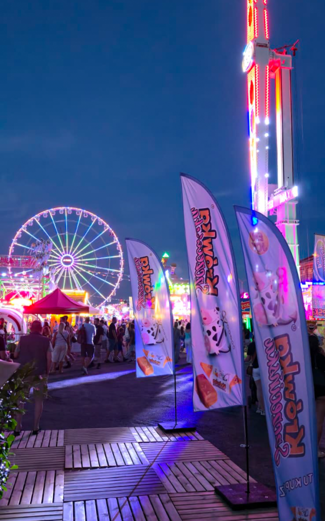 Nocny widok na flagi Krówki Szczecińskiej oraz rozświetlony lunapark i koło widokowe Wheel of Szczecin.